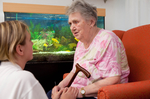 Eine Bewohnerin sitzt in einem Sessel vor einem Aquarium, eine Pflegerin hockt vor ihr und redet mit ihr.