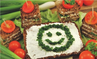 Verschiedene Scheiben Vollkornbrot mit Tomaten, auf einer mit Butter bestrichenen Scheibe ist mit grünen gehackten Kräutern ein lächelndes Gesicht abgebildet