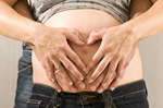 Aufnahme des Bauches einer Schwangeren. Hinter ihr steht ein Mann, beide haben ihre Hände auf ihren Bauch gelegt.